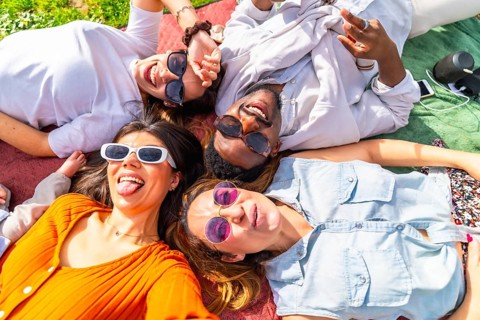 Four young multi ethnic friends are lying on a blanket, making funny faces and taking a selfie at a
