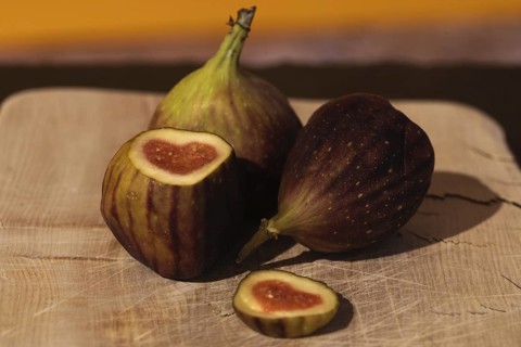 Three figs, one cut open, with a heart-shaped centre on a wooden board against a yellow background,
