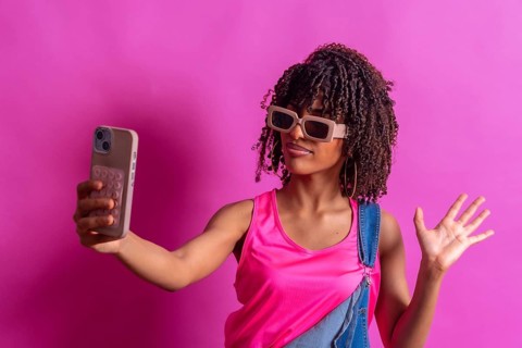Young woman wearing sunglasses and pink shirt taking selfie with her smartphone while waving with