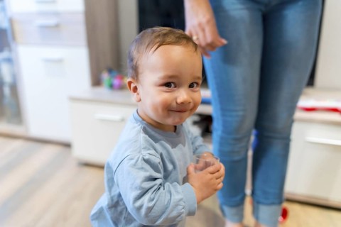 Happy toddler drinking water from a glass, standing close to his mother's legs in a cozy living