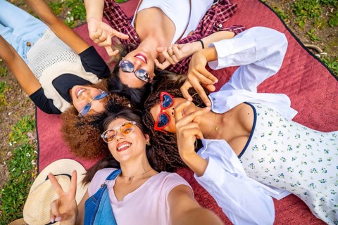 Four cheerful young women lying on a blanket in a park, making hand gestures and taking a selfie