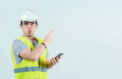 Surprised builder worker with phone pointing at blank space isolated. Amazed engineer holding phone