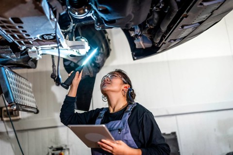 Young female mechanic using flashlight and digital tablet, inspecting underside of car raised on