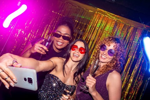 Three multi ethnic female friends are taking a selfie while enjoying sparkling wine at a nightclub,
