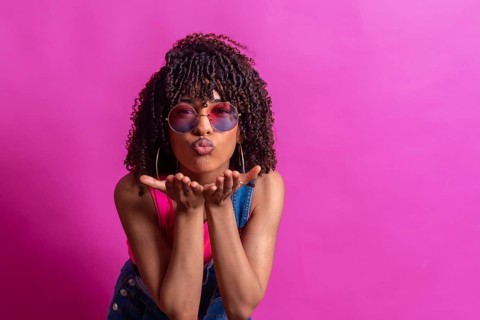 Fashionable young woman blowing a kiss while wearing sunglasses, set against a vibrant pink