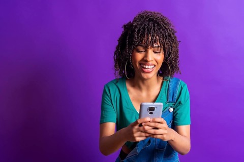 Young woman with curly hair, wearing casual clothes and smiling while using her smartphone against