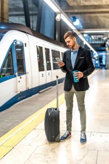 Businessman standing on subway platform, using smartphone, holding coffee, waiting for train with