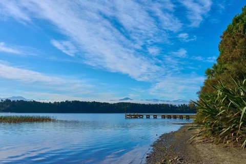 At the lake with pebble beach and plants, under a blue sky with clouds, with a wooden jetty, Lake
