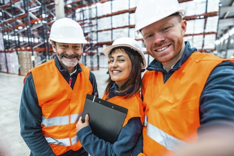 Warehouse workers wearing safety vests and hard hats are smiling and taking a selfie in a logistics