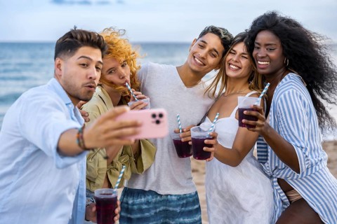 Group of cheerful multi ethnic friends taking selfie on beach while drinking juice and enjoying