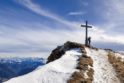 A hiker leans on the Hochgern summit cross with an impressive view over the Chiemgau mountains in