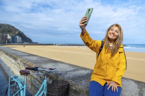 Young woman wearing yellow raincoat taking selfie with smartphone at la zurriola beach in san