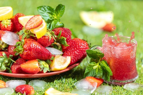 A sparkling drink next to strawberries, lemons, mint and ice cube cubes on grass