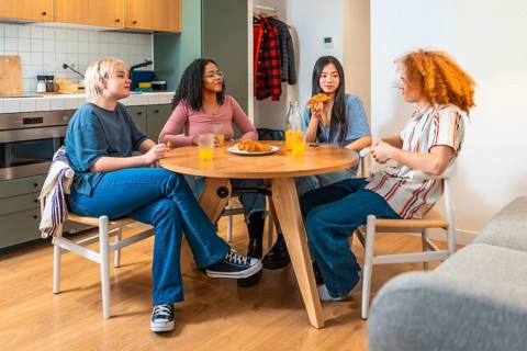 Four roommates are sitting around a wooden table in a modern kitchen, eating breakfast together and