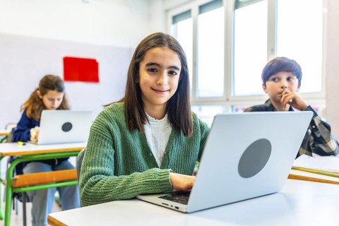 Elementary school student using laptop during class, smiling and looking at camera, with classmates