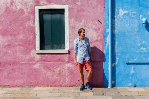 Young man leaning against a house wall, pink and blue house facade with window, colourful houses on
