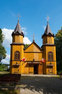 Front view of a wooden church with towers, Polish flags, surrounded by trees and blue sky, wooden