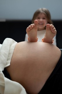 A pregnant woman with children's feet on her stomach, laughing child in the background, symbolic