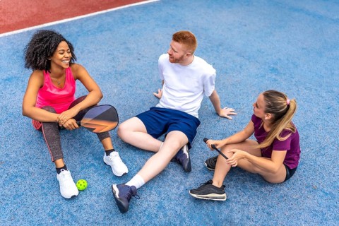 Top view photo of a multi-ethnic group of friends sitting on pickelball court chatting relaxed