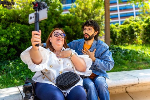 Two content creators sitting on a bench in a park, filming a video blog using a smartphone and