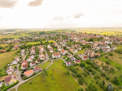 Densely built-up residential area with houses and gardens, surrounded by fields under a cloudy sky,