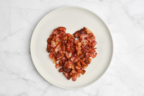 Crispy bacon pieces are creatively arranged on a white plate to form a heart shape, presenting an