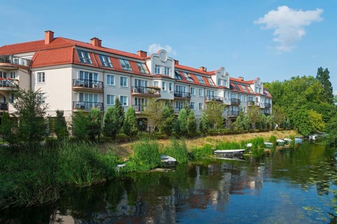 Modern residential buildings on a quiet riverbank with small boats and lush greenery in a summery