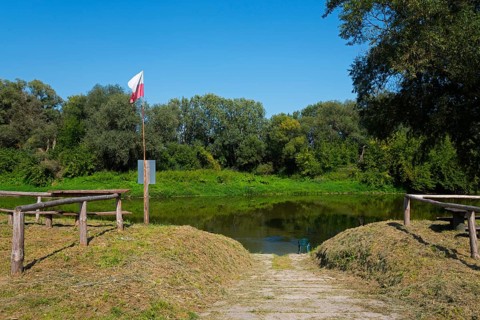 Riverbank with Polish flag and wooden railing, surrounded by green vegetation, bow River, border