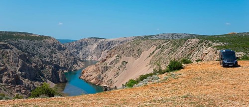 A motorhome stands on a rocky terrain overlooking a canyon with a river under a blue sky,