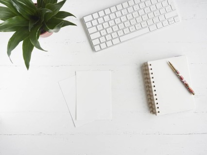 Notebook, blank sheets of paper and pen on white desk table beside a computer keyboard and a green