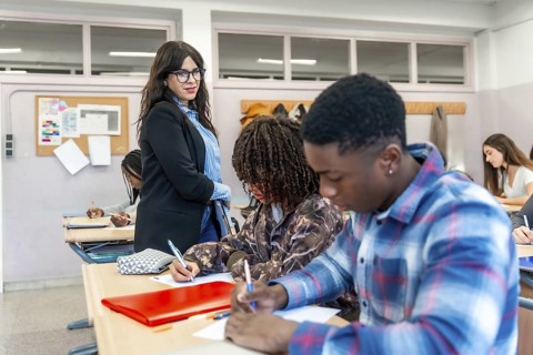 Multi ethnic high school students focused on writing exams in a classroom, with a teacher