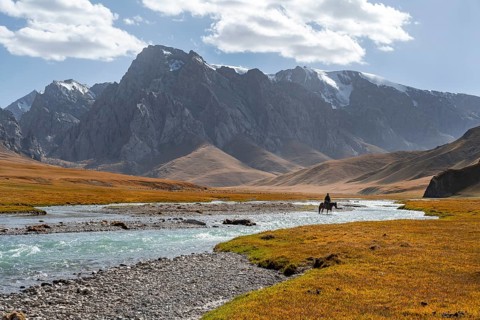 Rider riding through river in front of mountain landscape with yellow meadows, mountain peaks,