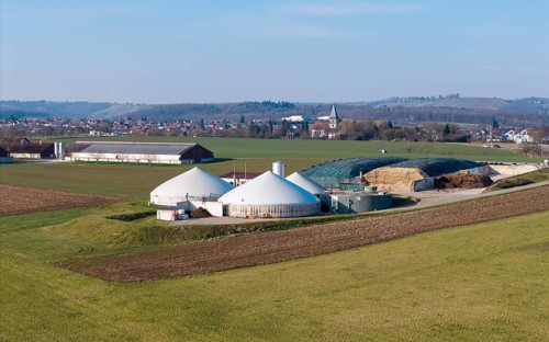 Biogas plant surrounded by fields, with buildings and village in the background on a slight hill,