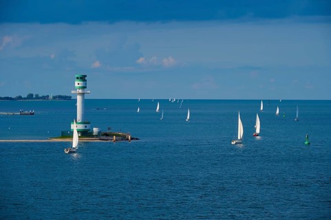 Several sailing boats and ships on calm blue sea under a cloudy sky with a lighthouse in the