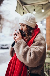 A young thoughtful woman sips a warm beverage outdoors in winter, wearing a white beanie and a cozy