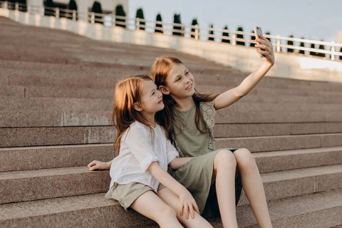 Two young girls sit on outdoor steps, smiling as they take a selfie with a smartphone. The sunny