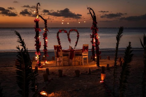 A romantic dinner table for two set on a beach at sunset with flower arches and decorative lights.