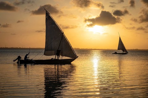 Two traditional sailboats are silhouetted against a serene, golden sunset on calm, reflective water