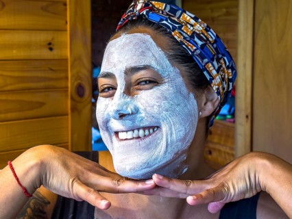 A woman smiling with a white face mask applied, posing playfully in a cozy wooden cabin in