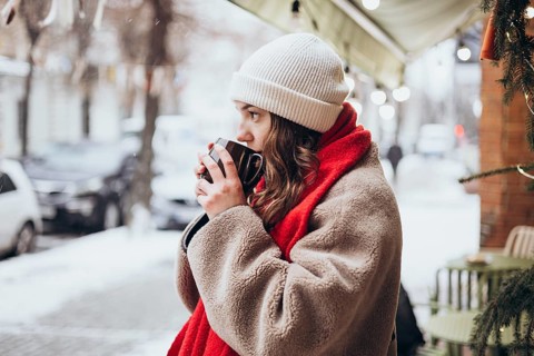 A young thoughtful woman is pictured standing outdoors on a snowy winter day, sipping from a mug.