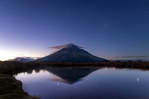Majestic Mount Taranaki stands under a starry sky, its peak enveloped in a gentle cloud. The still