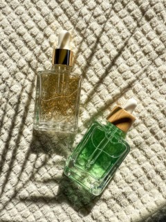 Two elegant dropper cosmetic bottles with gold and green serums lie on textured white fabric int he