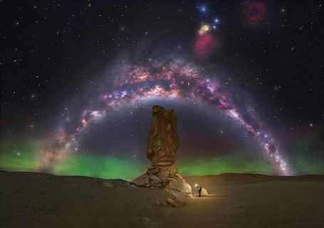 A lone figure stands beneath a towering rock formation, illuminated by the colorful arc of the
