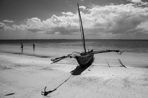 Black and white photograph of a traditional wooden dhow sailboat anchored on a sandy beach under a