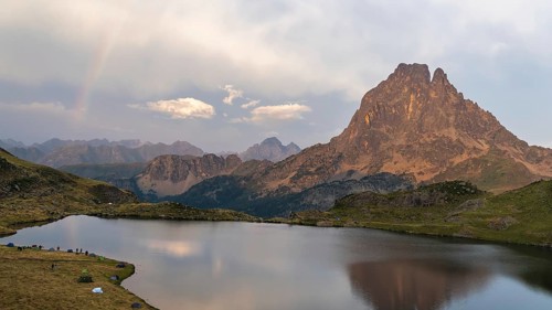 Panoramic view of the Midi d'Ossau peak reflected in a calm lake, surrounded by lush vegetation in