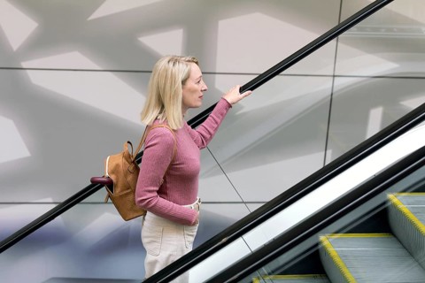 Woman rides an escalator in a supermarket, hand on railing, wearing a pink sweater and a brown