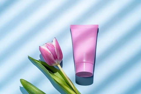 Top view of a pink cosmetic tube alongside a pink tulip lying on a blue background with light