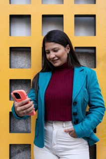 A latin woman wearing a teal blazer and burgundy sweater smiles while looking at her smartphone.