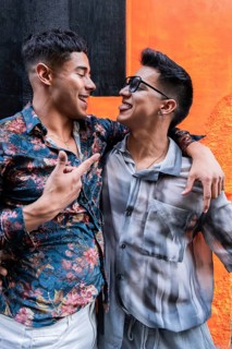 A cheerful latin gay couple shares a playful moment, smiling at each other against a vibrant orange