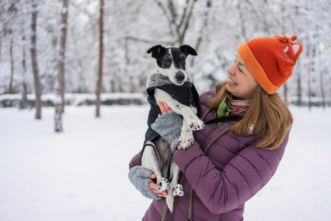 A cheerful woman in a bright hat and warm clothing holds her dog in a snow-covered park. The winter